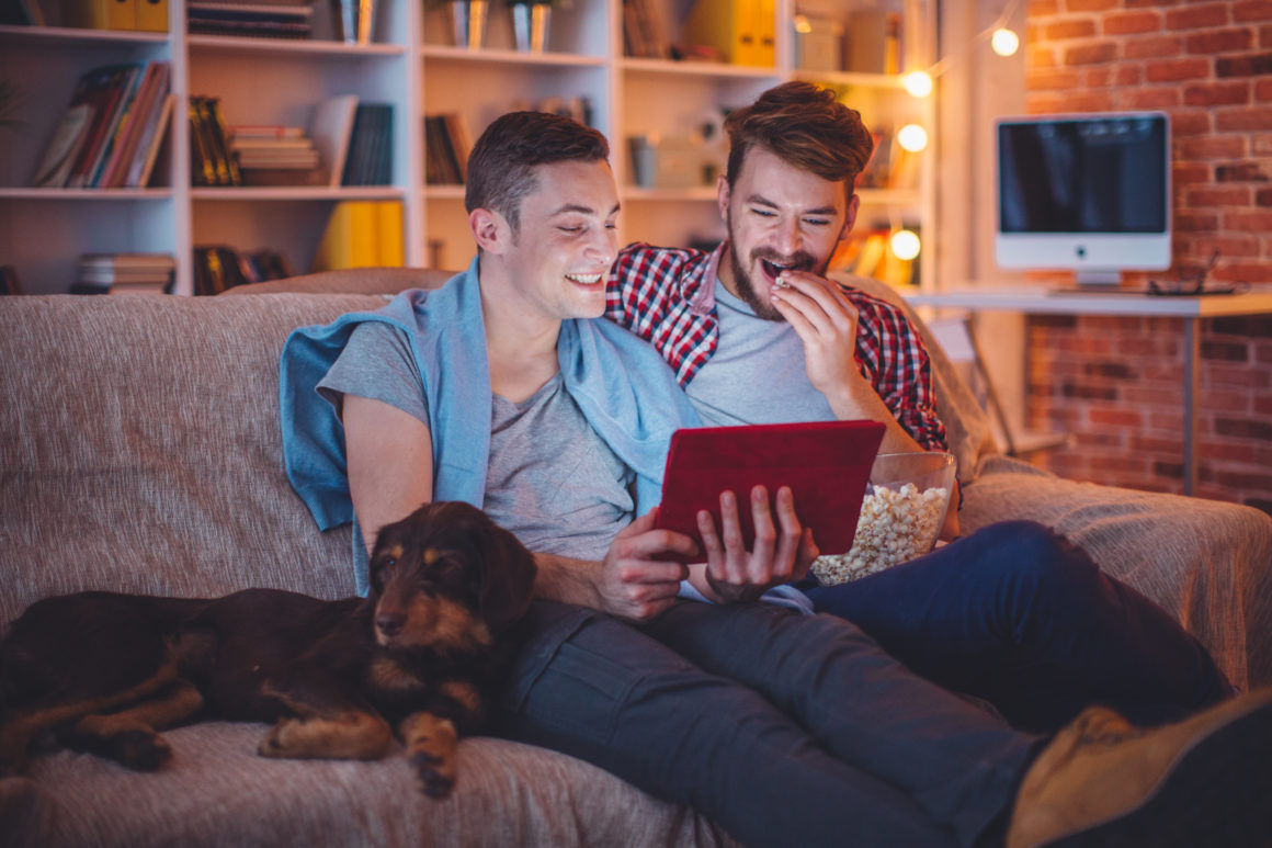 Young gay couple sitting on sofa at home having a movie night with their dog. Caucasian ethnicity, blond hair, casual.