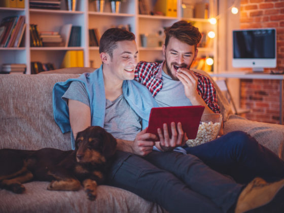Young gay couple sitting on sofa at home having a movie night with their dog. Caucasian ethnicity, blond hair, casual.